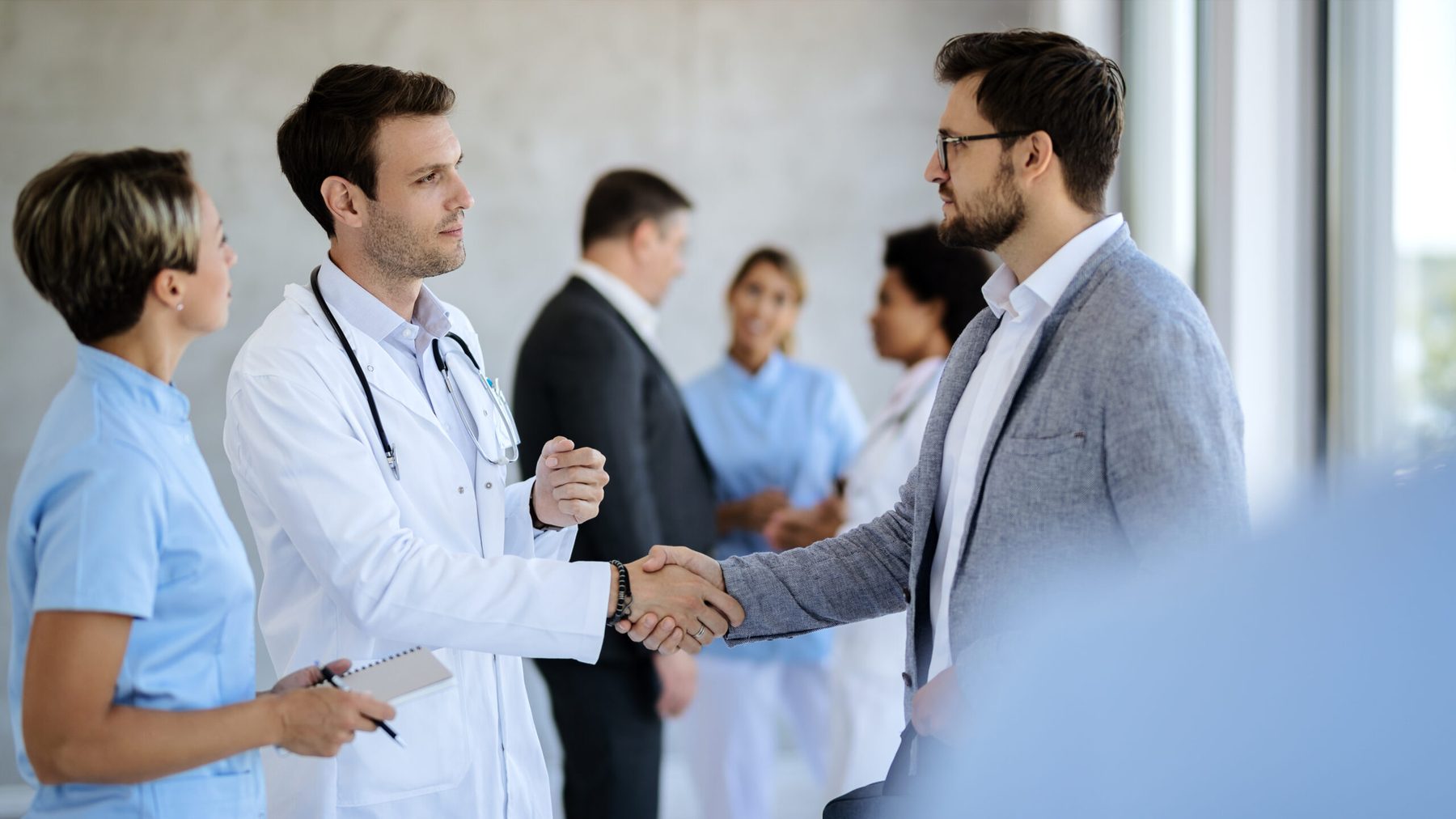 A doctor and a visitor in a jacket shaking hands in a hospital corridor while colleagues talk behind them