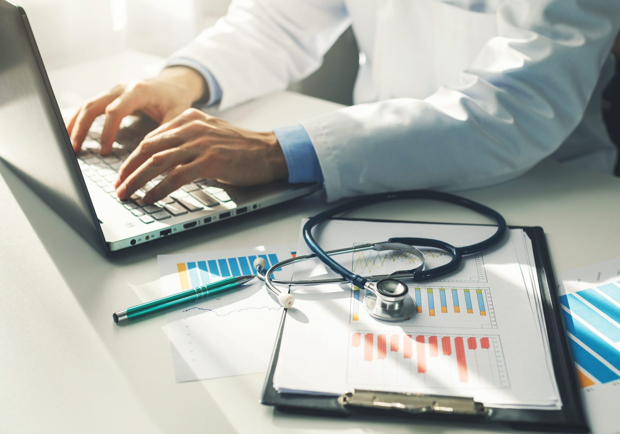 A clinician in a white coat typing on a laptop, printed charts and a stethoscope on the desk beside it